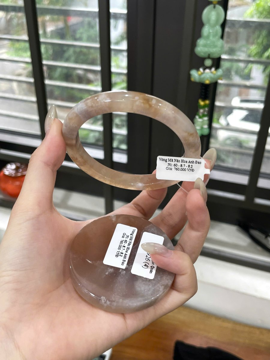 Vòng Mã Não Hoa Anh Đào Ni 60 ở phía trên bàn tay và vật tròn có tem ở bên dưới